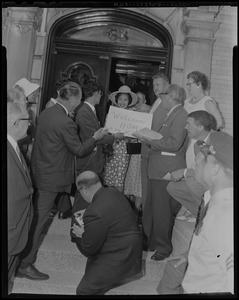 Queen Sirikit of Thailand standing by entrance to a building and holding a "Welcome Home to the King" sign