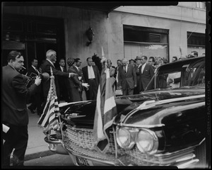 King Bhumibol Adulyadej and Queen Sirikit of Thailand on sidewalk with a crowd of people in front of automobile with diplomatic flags