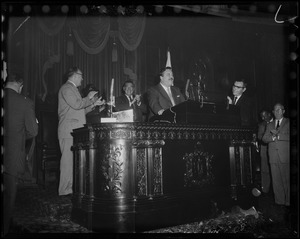 Filmstage-television comedian Jackie Gleason stands with other men at a speaker's podium as he addressed the House of Representatives at the State House