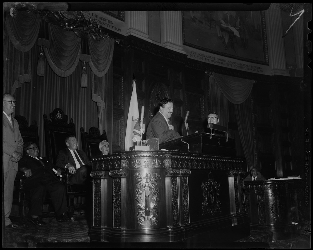 Filmstage-television comedian Jackie Gleason stands with other men at a speaker's podium as he addressed the House of Representatives at the State House