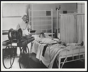 Peter Bent Brigham Hospital. 1913 - Picture of bedside unit of the first electrocardiograph machine in New England. Nurse is in touch with the laboratory in another part of the hospital. Cable on cart leads to lab. Electrical connections are put on patients leg.