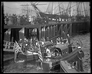 A group of people getting on boats off a pier, the USS Constitution in the background