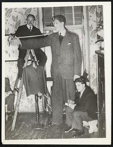 Job For A Tentmaker. Clambering around on a ladder a tailor is shown taking the measurements of Baine Myllyrinne, 8½-foot Finnish giant, when he dropped into a Vienna Tailor Shop recently to get a spring outfit. He needs 103 feet of cloth for a suit.