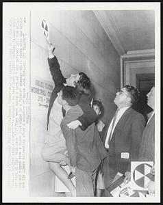 Dr. Thomas Brewer (wearing glasses) looks on as his wife Susan, is hoisted atop a companion's shoulder to tear down a civil defense fallout shelter sign at City Hall here 1/29. They were among a band of 9 who marched into the building and ripped down a dozen such signs in a "peace" rampage which ended with one arrest and the others retreating after a scolding by Mayor John Shelley.