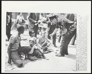 New York – A Word In Brooklyn – A patrolman leans over to talk to Mrs. Irene Parker, 23, mother of Godfrey, 4, wearing paper hat, as they sat with companions on a street at a Brooklyn construction site. Others in line are Claude Jones, 13, left, and Deborah Poore, 11, right. The patrolman told Mrs. Parker she was violating the law and would have to move. Minutes later a shoving and pushing session started in which Mrs. Parker and her son were involved.