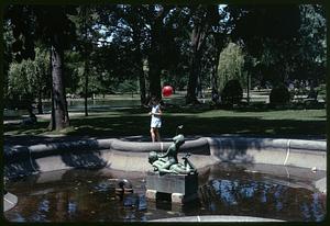 A person holding a red balloon walking past Triton Babies fountain, Boston Public Garden