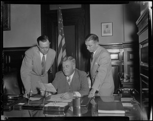 Man seated at desk reading, while two others look on