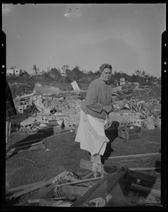 Woman wearing an apron, standing with folded hands next to destruction left by tornado