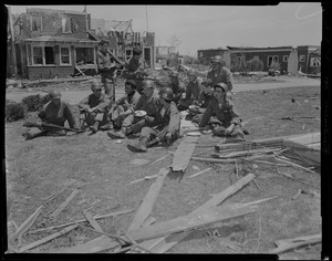 Group of military personnel standing and sitting, with buildings damaged by tornado behind them