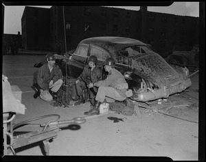 Military personnel seated beside a damaged car, one holding a telephone receiver