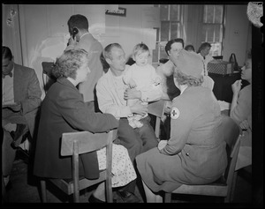 Group of people seated, including man holding a child and possibly Red Cross personnel