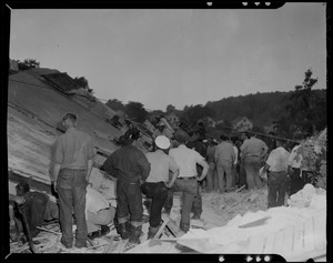 Group of people, including emergency personnel, standing amid wreckage, most with their backs to the camera