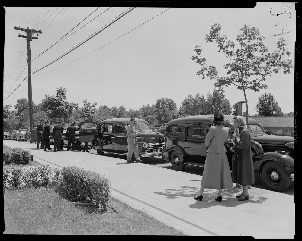 Pallbearers loading coffin into hearse, with bystanders and other cars nearby