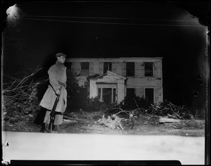 Military man standing with rifle in front of downed trees and building damaged by tornado