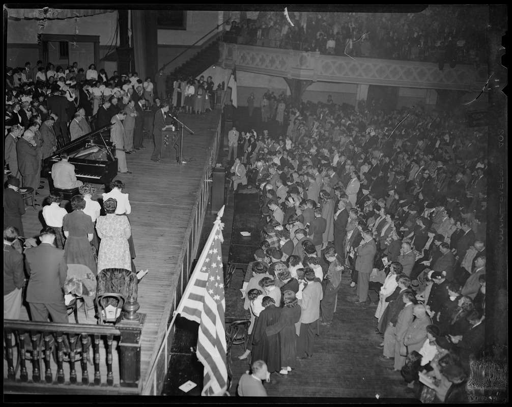 Large crowd praying at revival meeting at Mechanics Hall, with Billy Graham speaking from podium