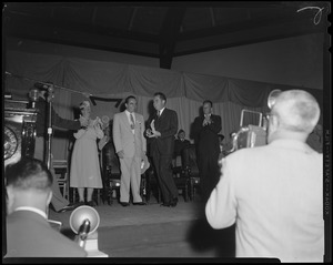 Group of people standing on stage while photographers take pictures, including Billy Graham applauding on the right
