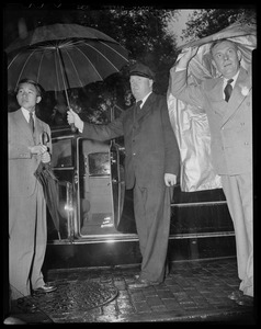 Crown Prince Akihito standing next to open door of vehicle with man holding umbrella open and other man covering head with coat