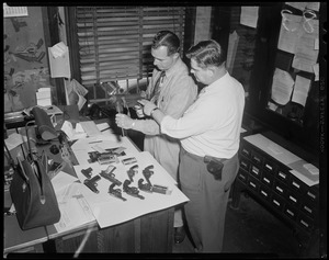 Found in raid -- cache -- Roger White (left) and Frank Bailey, both ballistics men examine sub-machine gun at Boston Police Headquarters after it was confiscated in a Back Bay lodging house along with five loaded revolvers (on table), following a raid by detectives