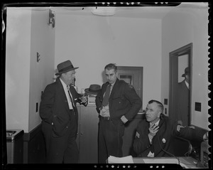 Two men standing together next to police sergeant seated at desk, on telephone