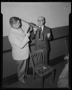 Two men examining gun, next to chair holding various handguns