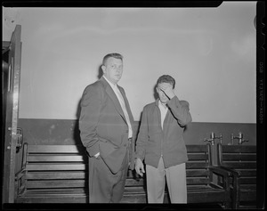 Man standing, handcuffed to Elmer "Trigger" Burke, who is covering his face with left hand