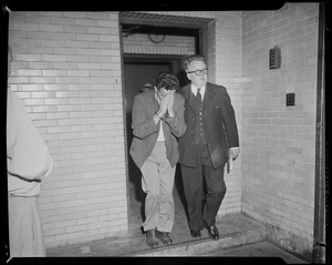 Man escorting Elmer Burke who is handcuffed and hiding his face behind his hands