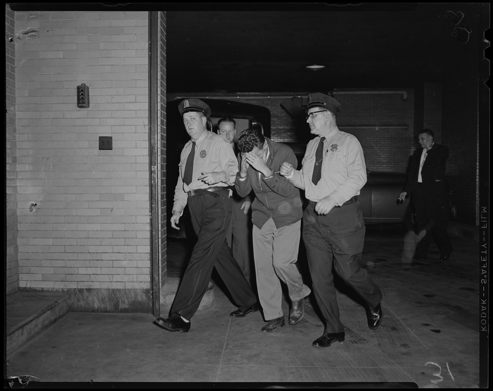 Two police officers, both handcuffed to Elmer Burke, who is covering his face with his hands