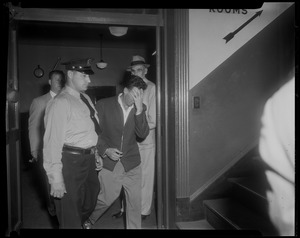 Elmer Burke, handcuffed to police officer, covering face with left hand near stairs