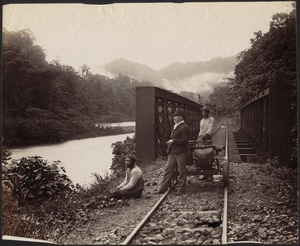 Three men standing on tracks near railroad bridge looking out over river; hand-pulled cart on tracks with luggage; misty mountains in distance.