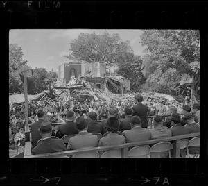 Crowd of people seating on chairs in rows listening to man speaking with destroyed building in background