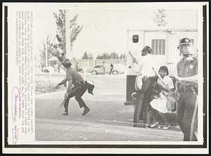 Police Throw Rocks Too -- A Dade County sheriff's deputy steps from behind a police van to throw rocks at Negroes in the Violence today at Florida Memorial College. Two policemen at right stay under cover as rocks from the Negroes pour in. The woman with the gas mask accompanied the police in their battle with the students.