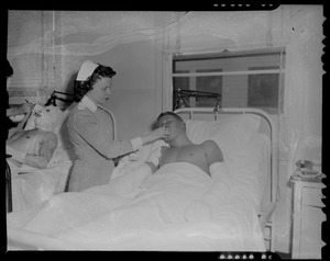 An injured person lying in a hospital bed being tended to by a nurse, likely military personnel injured in the U.S.S. Bennington aircraft carrier explosion off coast of Rhode Island