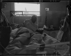 An injured person lying in a hospital bed speaking with a journalist, likely military personnel injured in the U.S.S. Bennington aircraft carrier explosion off coast of Rhode Island