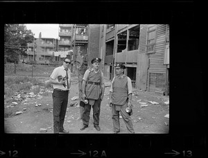Man with movie camera and two policeman, wearing bullet-proof vests, keep a surveillance in the rear of a Dudley St., Roxbury, building