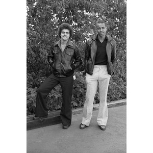 Full-length portrait of two Hispanic men wearing leather jackets, standing, facing front, at a Latino street festival