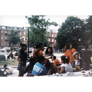 Teenagers in blue and orange staff shirts cluster around a table in the playground.