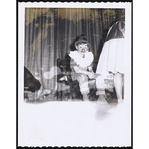 A boy wearing a clown costume sits in a chair on the stage for a Boys' Club's costume contest