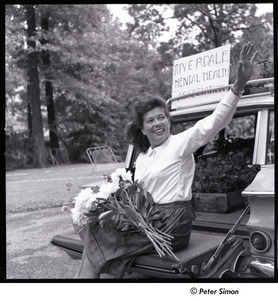 Andrea Simon: sitting on car trunk, waving, holding flowers in front of sign that reads, 'Riverdale Mental Health Association'
