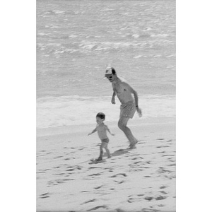 Unidentified man running behind a toddler on the sand at the beach.