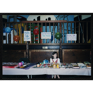 A girls sits behind table arranged with new toys. Sings posted above the table read (l to r) "Give Peace A Chance", "Toy Gun Buyback Today 4:00-6:00 Trade in your Toy Guns for a new toy!!!" and "You must trade in a toy gun or toy weapon to get one of the new toys on the table!"