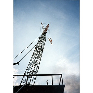 View from below of a crane lowering a steel beam bearing an American flag onto the platform of the Taino Tower construction site.
