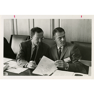 Richard Harte, Jr., looks to his left while turning a page on a document as an unidentified man looks on at a Boys' Club meeting