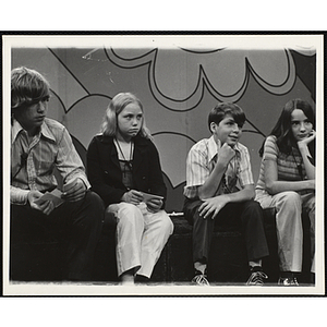 Four teenage girls and boys wear microphones around their necks and sit together on the set of a television show [?]