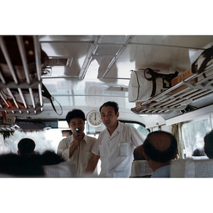 Men stand at the front of a tour bus, one speaking to passengers through a microphone