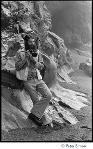 Rameshwar Das: full-length portrait with camera against an eroded granite outcropping on the beach