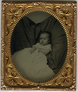 Portrait of unidentified infant in ample gown, lying on a striped pillow