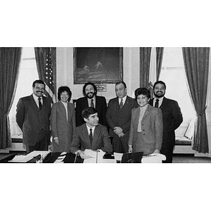 Michael Dukakis, seated at desk, with six Hispanic American leaders standing behind him, including Nelson Merced (left), at a political event in the Governor's office