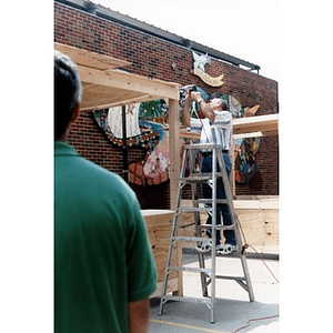 Man on a ladder screwing together the plywood frame of a concession stand for Festival Betances.