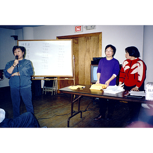 Woman speaks into the microphone discussing Association elections, with a ballot box and a whiteboard showing names of canditates visible to the right