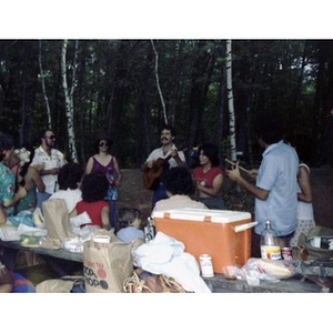 Approximately fourteen people play musical instruments, sing, and clap their hands by a table covered with food, at a La Alianza staff picnic at an unidentified location in the woods.